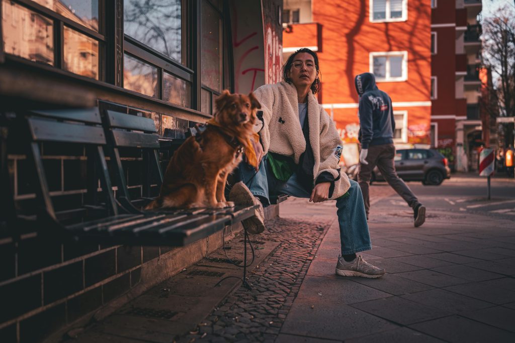 Chaotic street scene in Berlin featuring a person and a dog sitting on a bench with a passerby in the background.